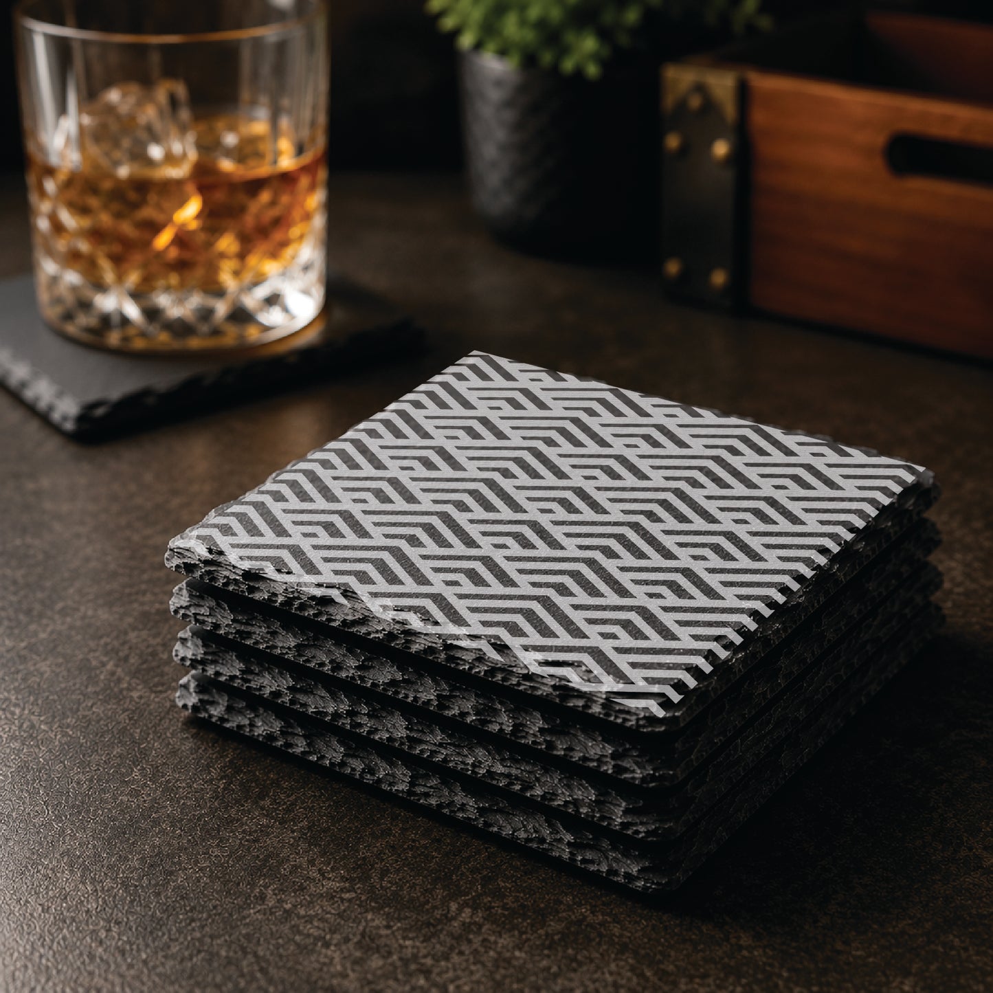 Stacked set of Geometric Peaks engraved slate coasters on a dark tabletop with whiskey glass and bar décor.