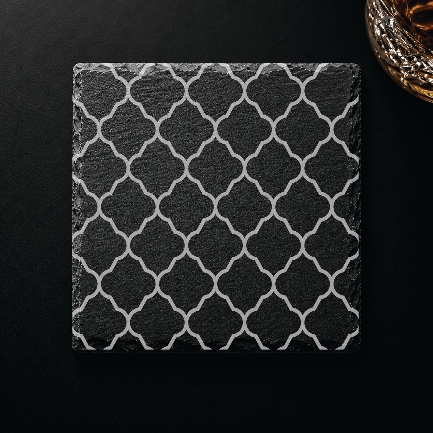 Quatrefoil engraved slate coaster displayed on a dark tabletop beside a whiskey glass, highlighting an elegant geometric lattice design.