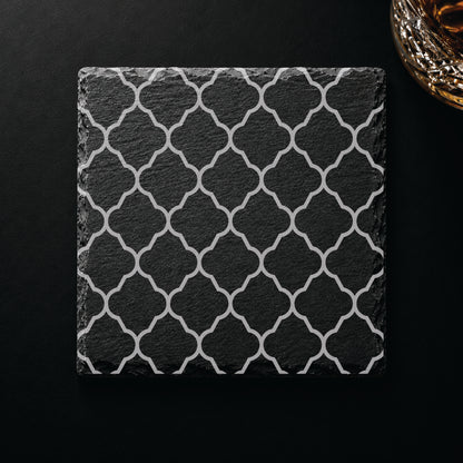 Quatrefoil engraved slate coaster displayed on a dark tabletop beside a whiskey glass, highlighting an elegant geometric lattice design.