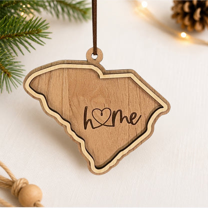 Layered South Carolina wood ornament featuring engraved “home” script and faux leather hanger on a soft neutral holiday background with greenery and warm lights.