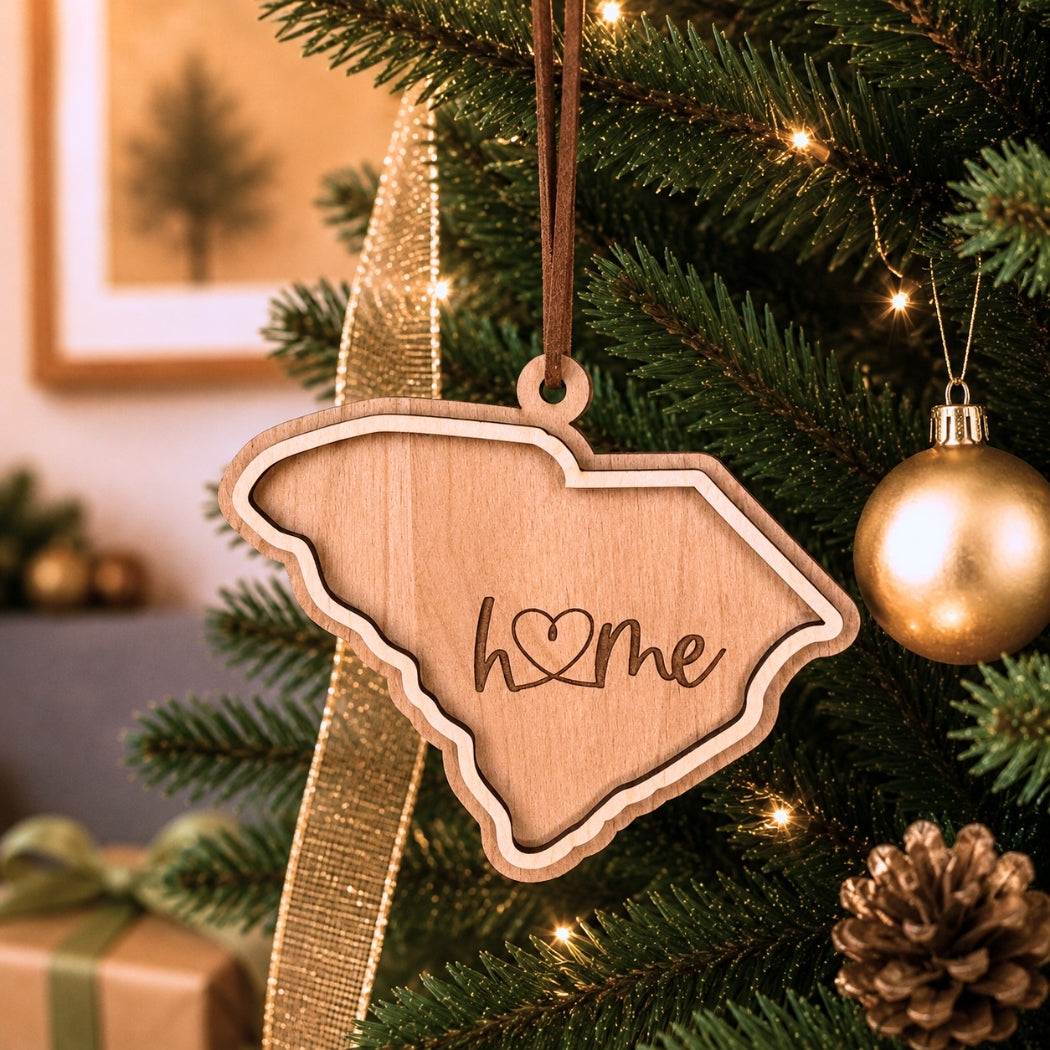 Layered South Carolina “home” ornament hanging on a decorated Christmas tree with warm white lights, pinecones, and gold ornaments.