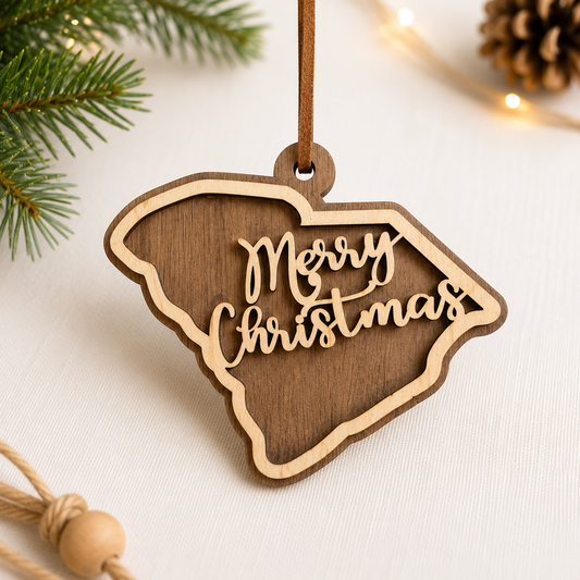 Layered hardwood South Carolina Christmas ornament featuring walnut and maple wood with “Merry Christmas” script detail and faux leather hanger styled with evergreen branches and warm holiday lights.