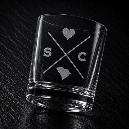 Angled engraved South Carolina whiskey glass featuring frosted South Carolina silhouette and modern crossed-letter design on a dark textured surface.