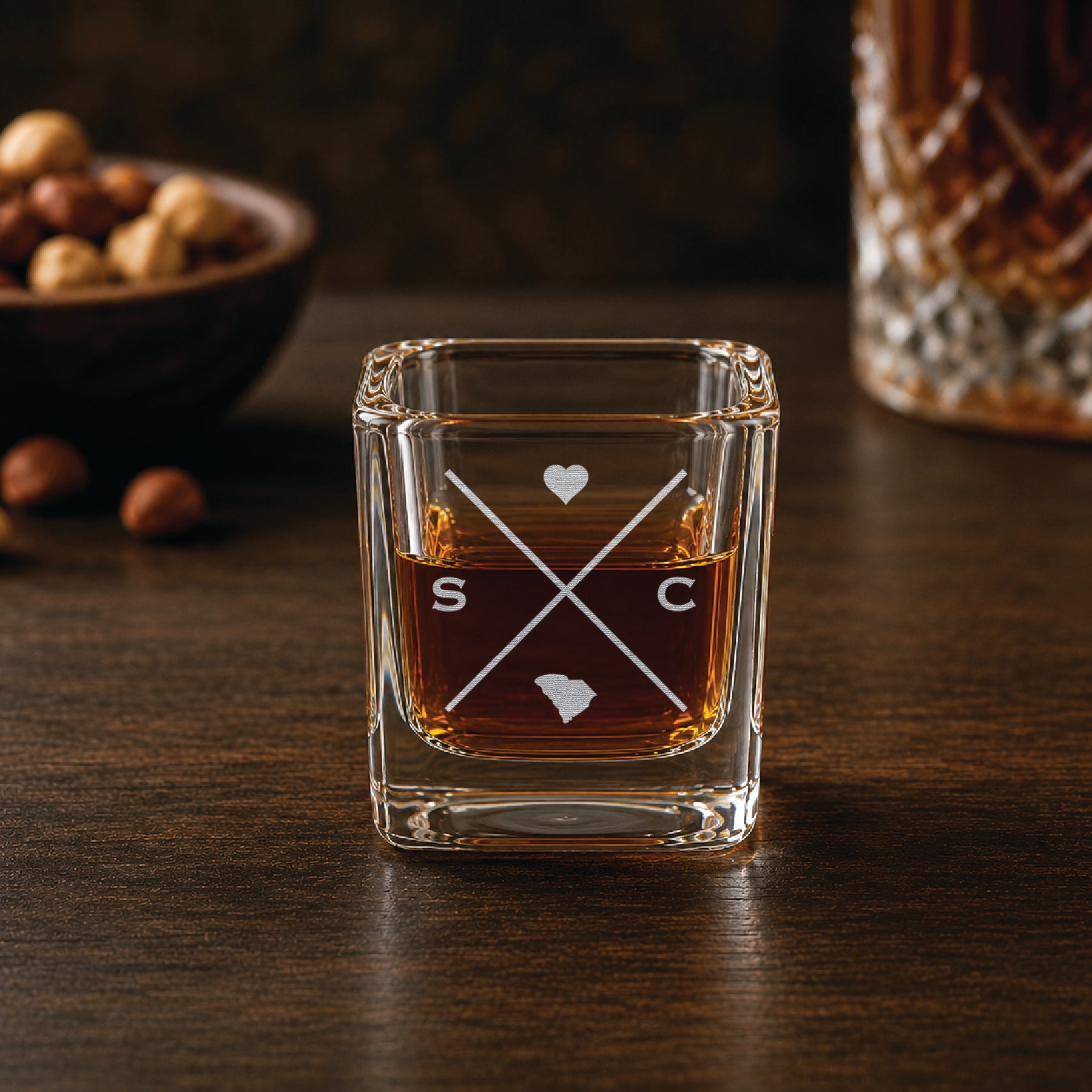 Engraved South Carolina shot glass filled with whiskey in a warm bar-style setting, featuring a minimalist crossed-line design with heart detail and South Carolina silhouette.