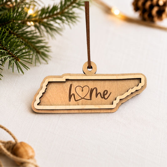 Layered Tennessee wood ornament featuring engraved “home” script and faux leather hanger on a soft neutral holiday background with greenery and warm lights.