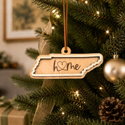 Layered Tennessee “home” ornament hanging on a decorated Christmas tree with warm white lights, pinecones, and gold ornaments.