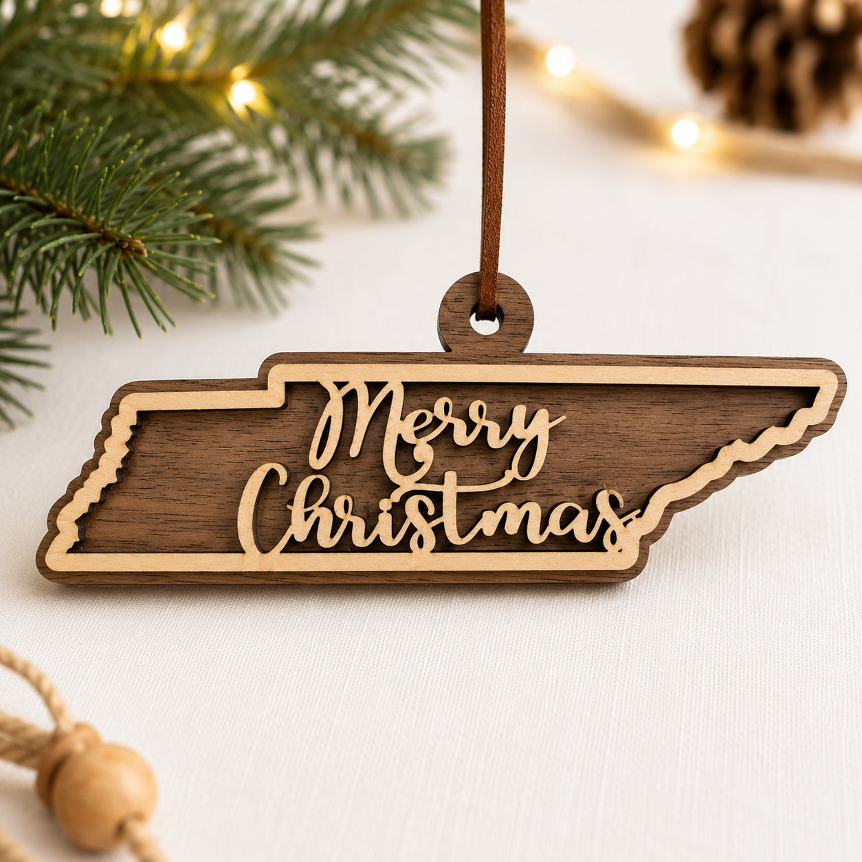 Layered hardwood Tennessee Christmas ornament featuring walnut and maple wood with “Merry Christmas” script detail and faux leather hanger styled with evergreen branches and warm holiday lights.