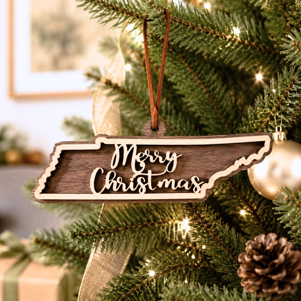Layered Tennessee state Christmas ornament hanging on a decorated holiday tree, featuring walnut and maple wood tones, “Merry Christmas” script, warm white lights, and neutral festive décor.