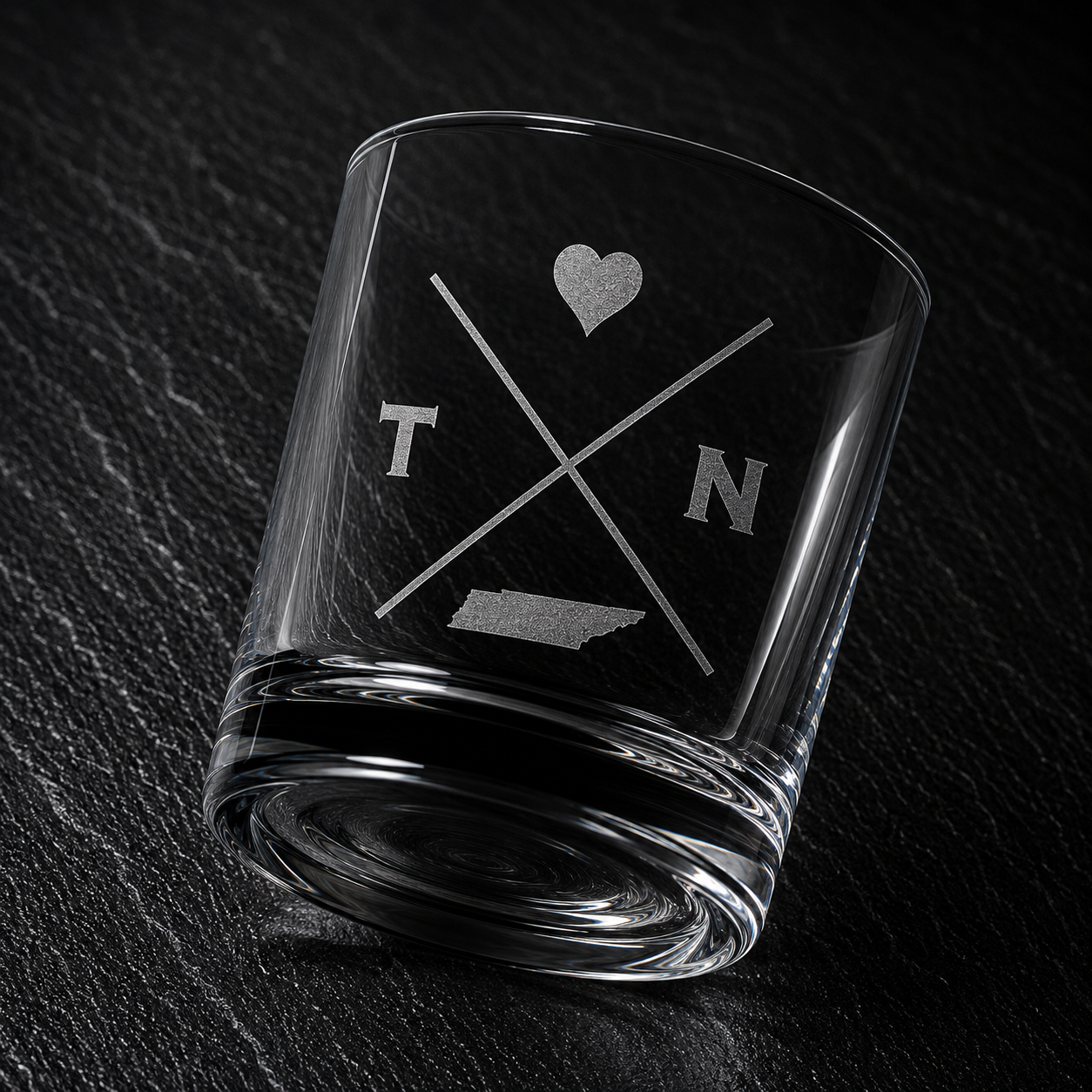 Angled engraved Tennessee whiskey glass featuring frosted Tennessee silhouette and modern crossed-letter design on a dark textured surface.