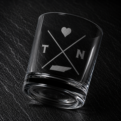 Angled engraved Tennessee whiskey glass featuring frosted Tennessee silhouette and modern crossed-letter design on a dark textured surface.