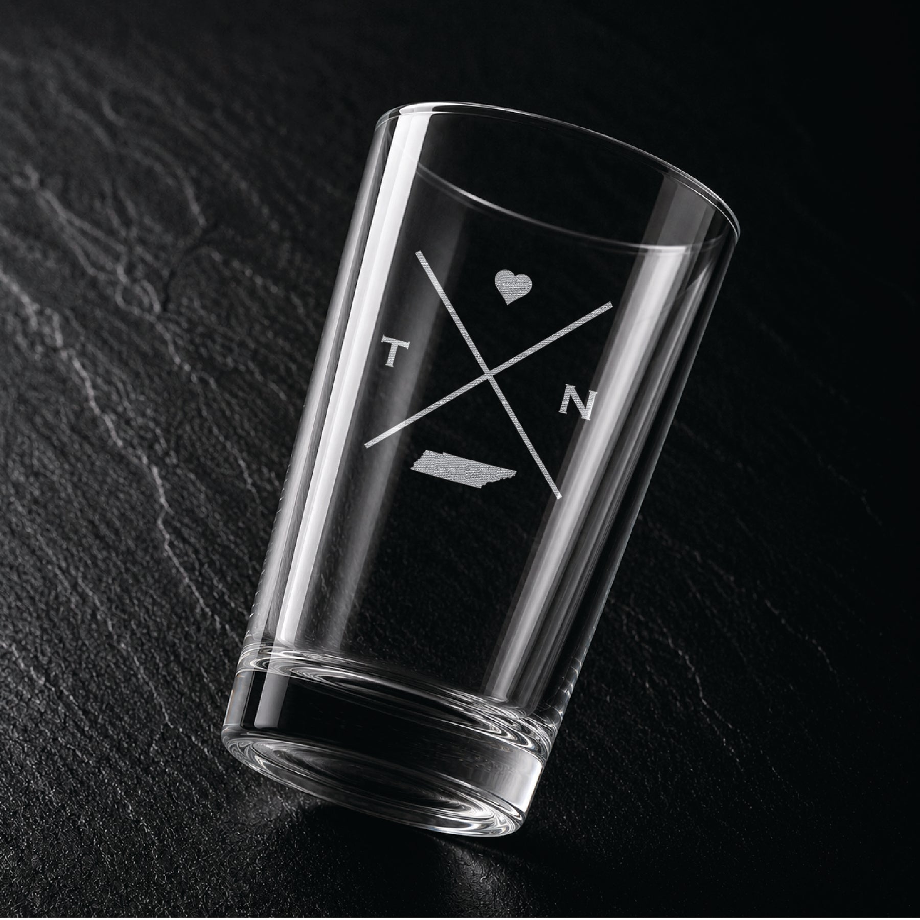 Angled view of engraved Tennessee pint glass with modern crossed-line state design and frosted laser engraving on dark textured surface.