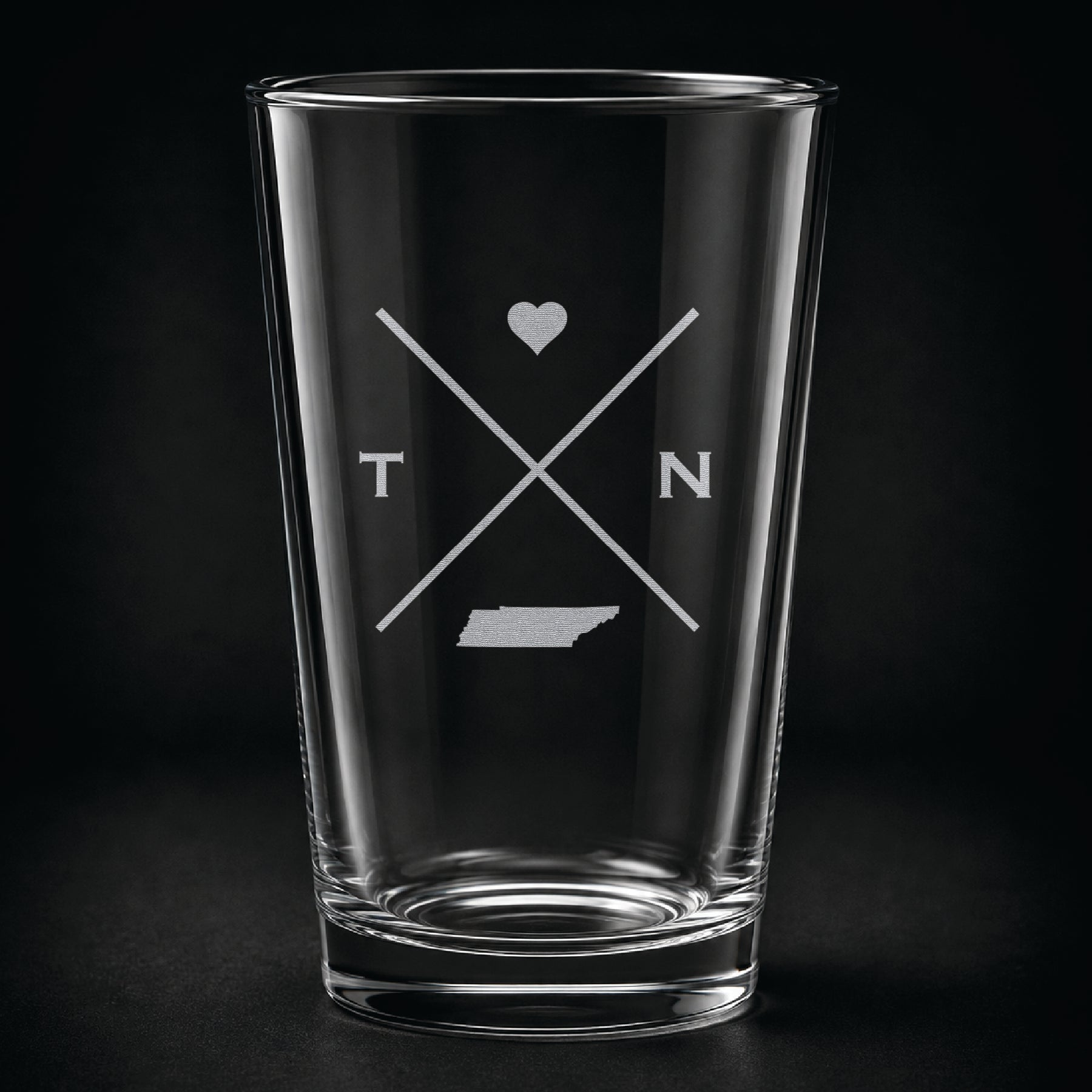 Engraved Tennessee pint glass featuring a minimalist crossed-line design with heart detail and Tennessee silhouette on a dark background.