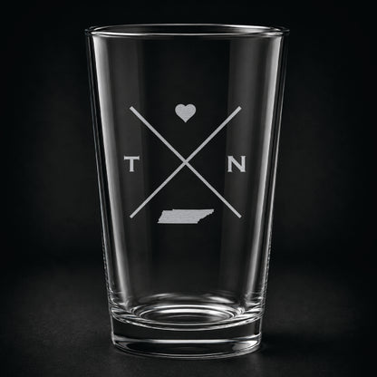 Engraved Tennessee pint glass featuring a minimalist crossed-line design with heart detail and Tennessee silhouette on a dark background.