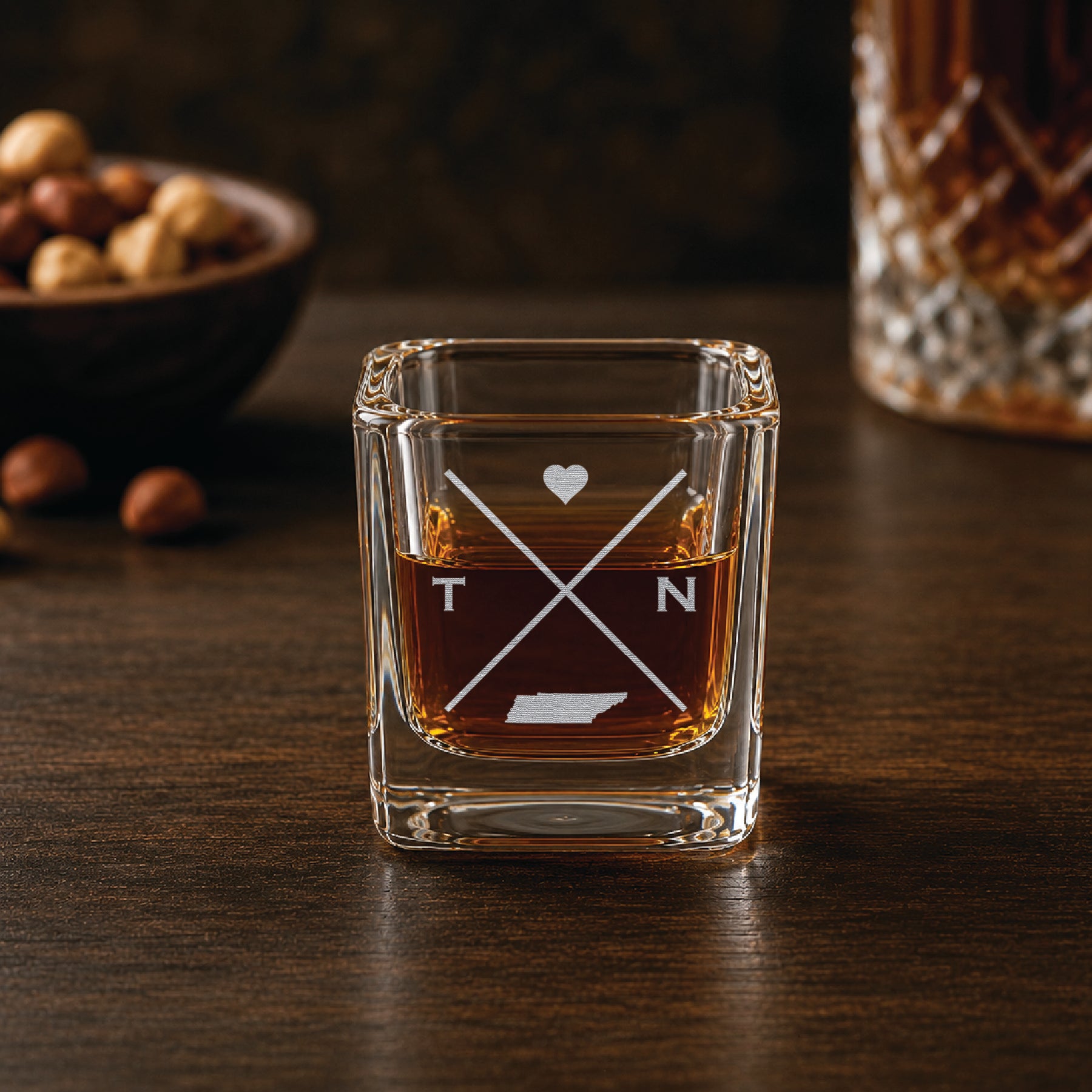 Engraved Tennessee shot glass filled with whiskey in a warm bar-style setting, featuring a minimalist crossed-line design with heart detail and Tennessee silhouette.