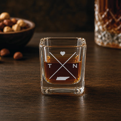 Engraved Tennessee shot glass filled with whiskey in a warm bar-style setting, featuring a minimalist crossed-line design with heart detail and Tennessee silhouette.
