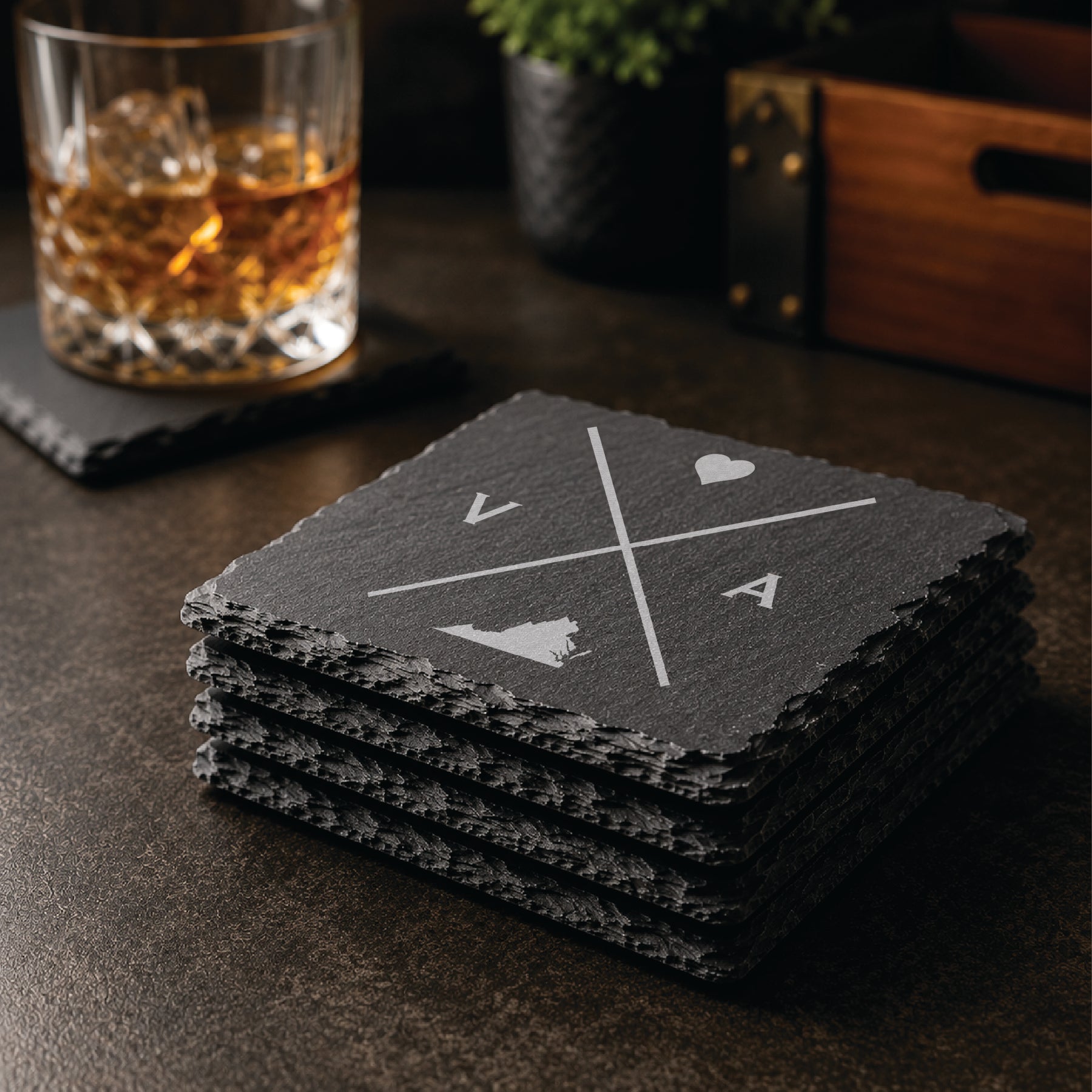 Stack of engraved Virginia slate coasters in a warm bar-style setting with whiskey glass nearby, featuring a minimalist crossed-line Virginia design.