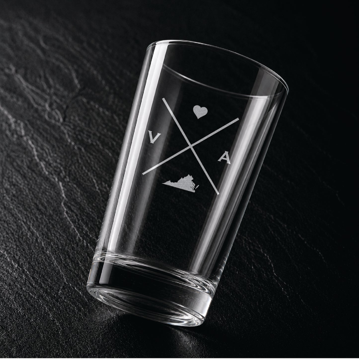 Angled view of engraved Virginia pint glass with modern crossed-line state design and frosted laser engraving on dark textured surface.
