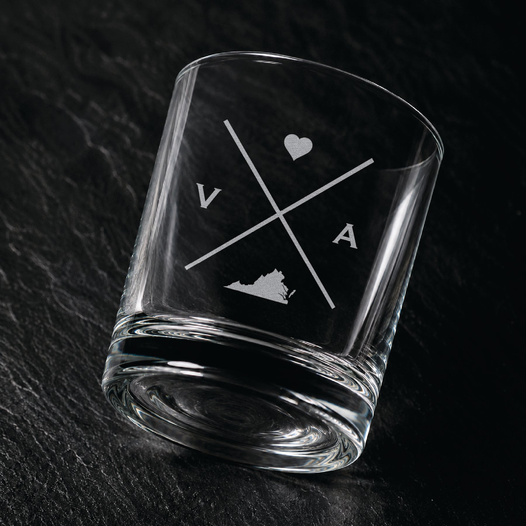 Angled engraved Virginia whiskey glass featuring frosted Virginia silhouette and modern crossed-letter design on a dark textured surface.