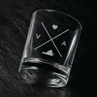 Angled engraved Virginia whiskey glass featuring frosted Virginia silhouette and modern crossed-letter design on a dark textured surface.