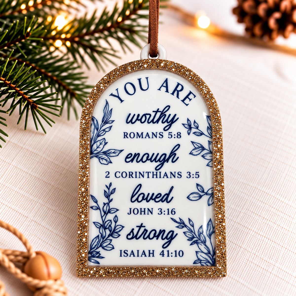 A Christmas ornament shaped like a gold glitter-framed arch with blue text reading 'YOU ARE worthy enough loved strong' and Bible verses, hanging on a light fabric surface with pine branches and warm string lights in the background.