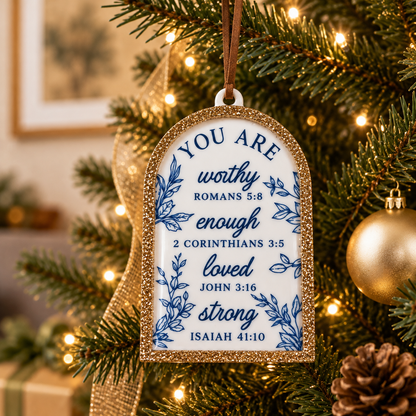A Christmas ornament shaped like a gift tag hangs on a decorated tree. The ornament has a white center with blue decorative text and floral illustrations, surrounded by a glittery gold border.