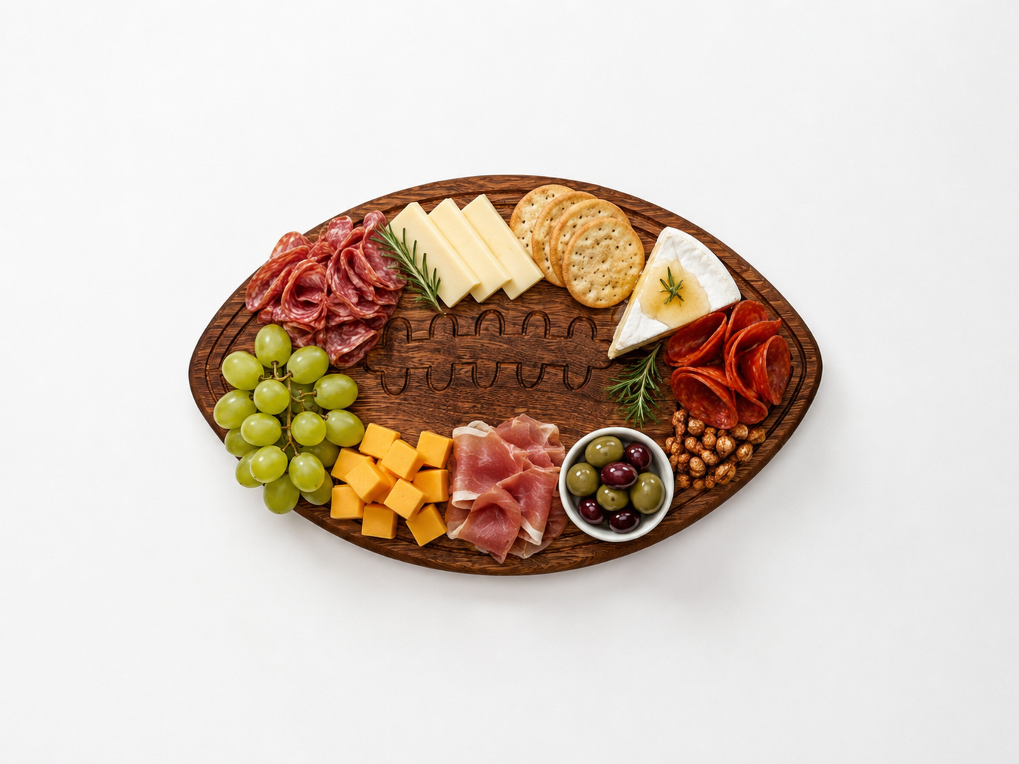 Football-shaped wooden charcuterie board with engraved laces, styled flat on a clean white background and filled with an assortment of cheeses, cured meats, crackers, grapes, olives, and nuts arranged neatly around the edges.